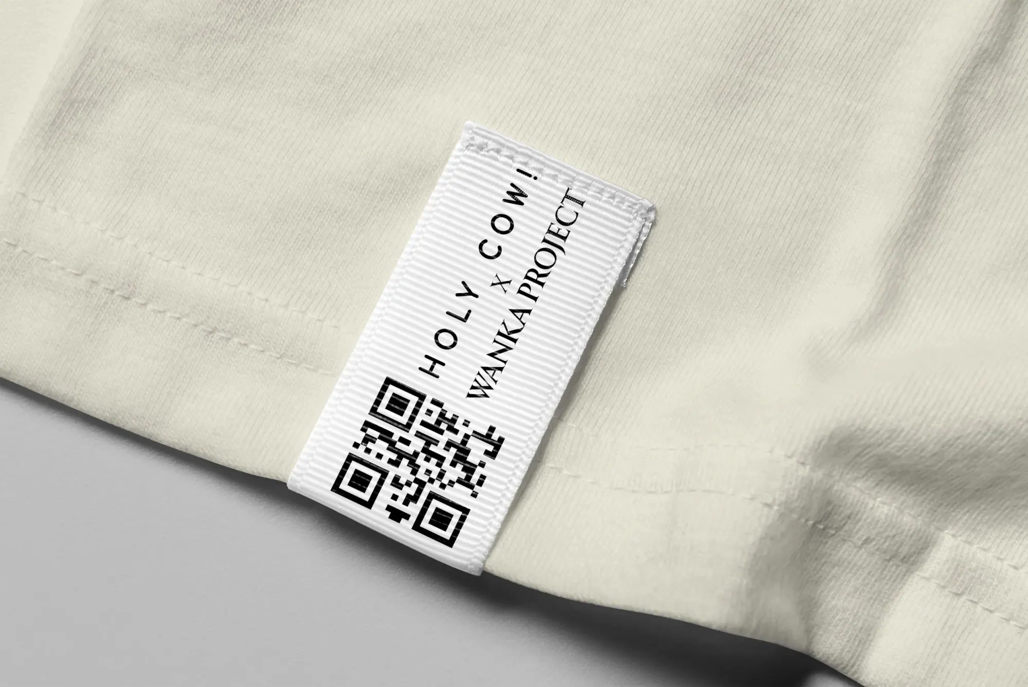 Étiquette de vêtement blanc avec code QR et texte 'HOLLY COW! WANKA PROJECT' sur un tissu beige.
