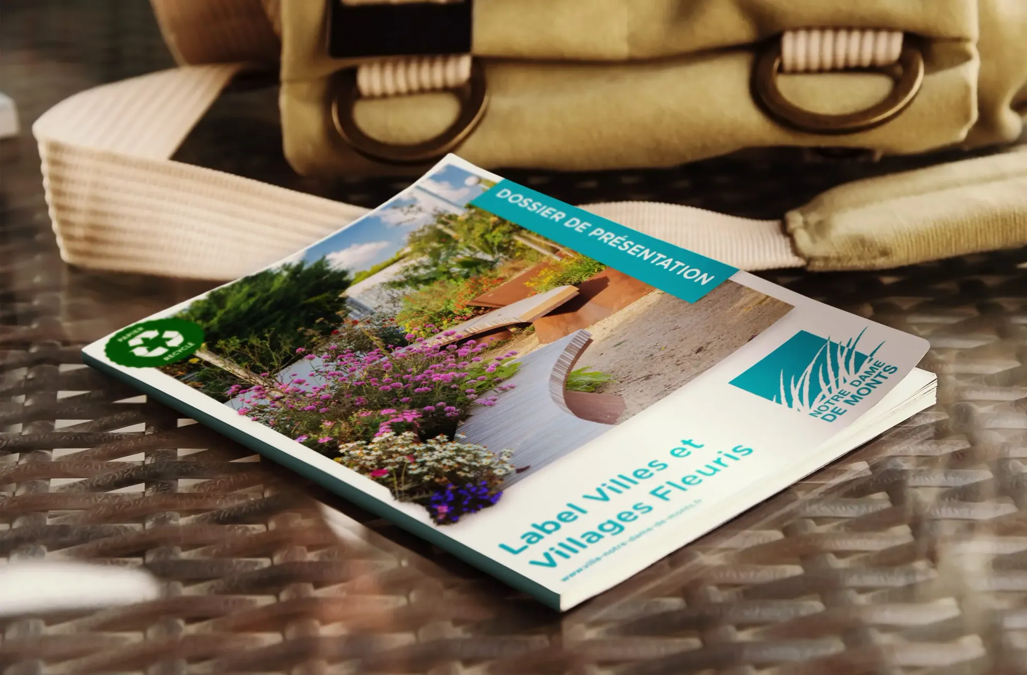 Brochure touristique mettant en avant la ville de Villages Fleuris, avec des fleurs colorées et un aménagement extérieur, placé sur une surface en rotin, à côté d'un sac beige.